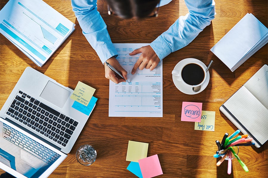 Person writing on a document with graphs, laptop, coffee cup, and sticky notes on a wooden desk, illustrating business writing and professional training context.