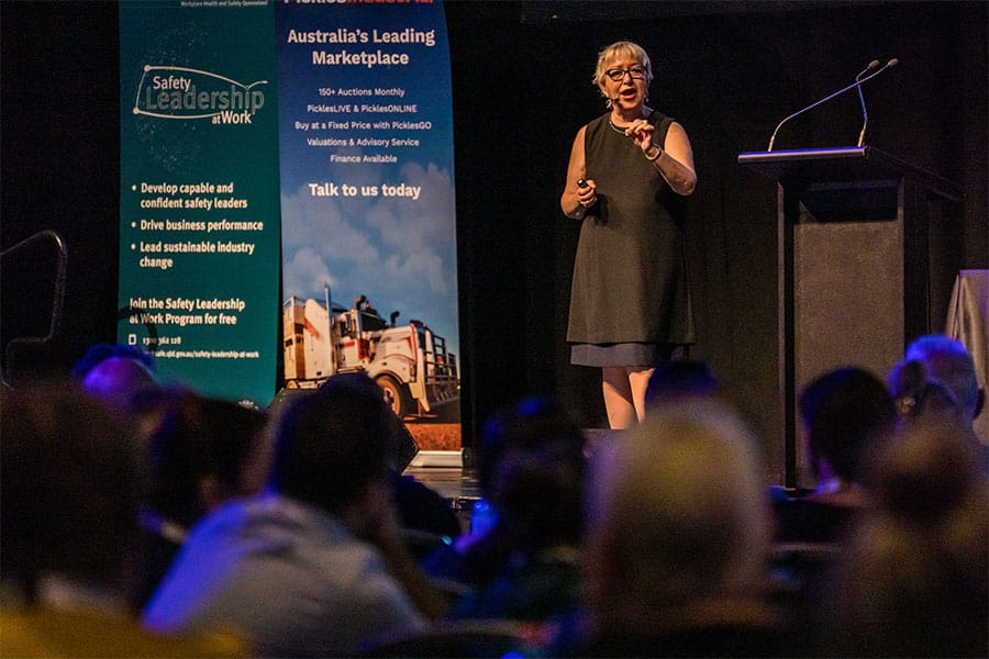 Woman presenting on stage at a professional training event with a focus on leadership and safety, audience visible in the foreground, banners promoting safety leadership programs in the background.