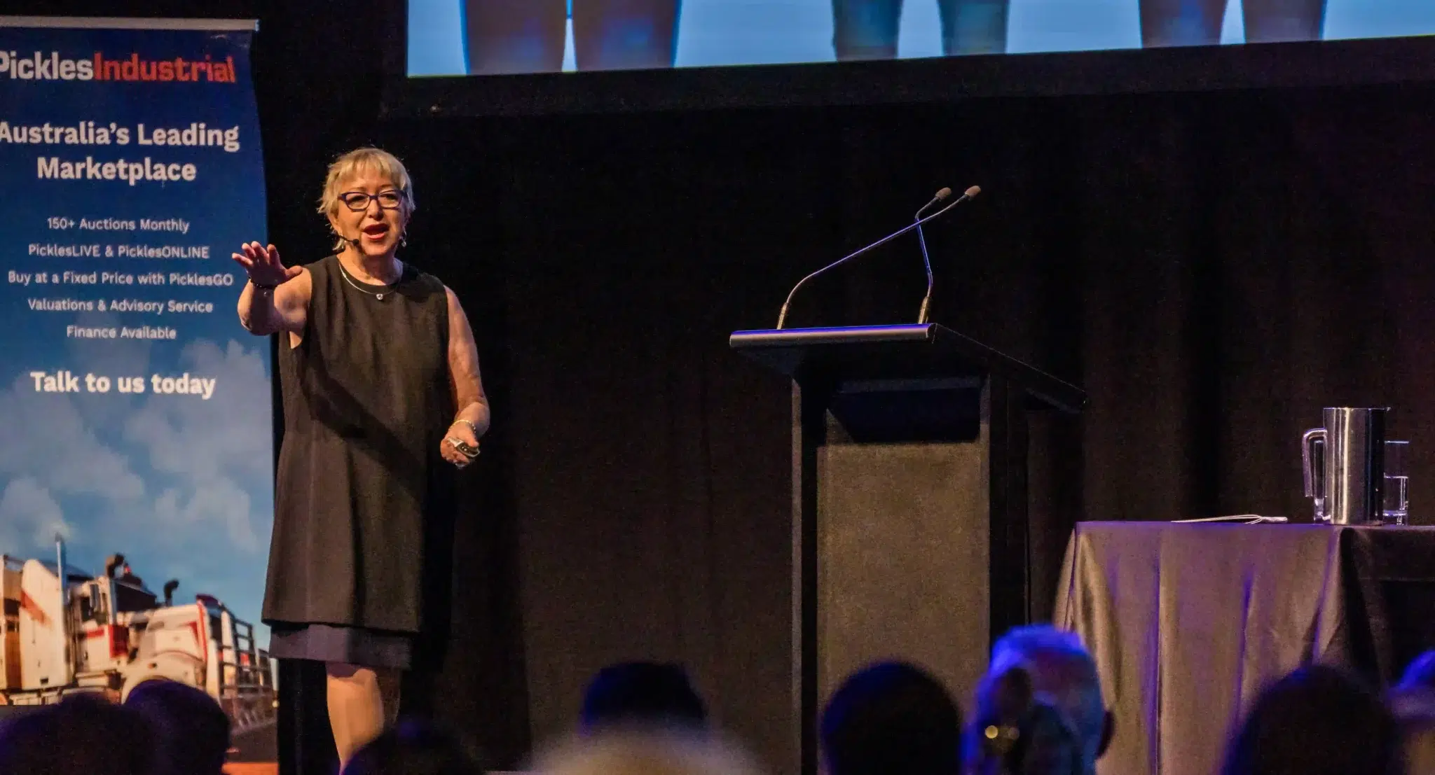 Nina Sunday presenting on stage, demonstrating effective communication skills, with a backdrop featuring promotional material for Pickles Industrial, highlighting her expertise in workplace training and development.